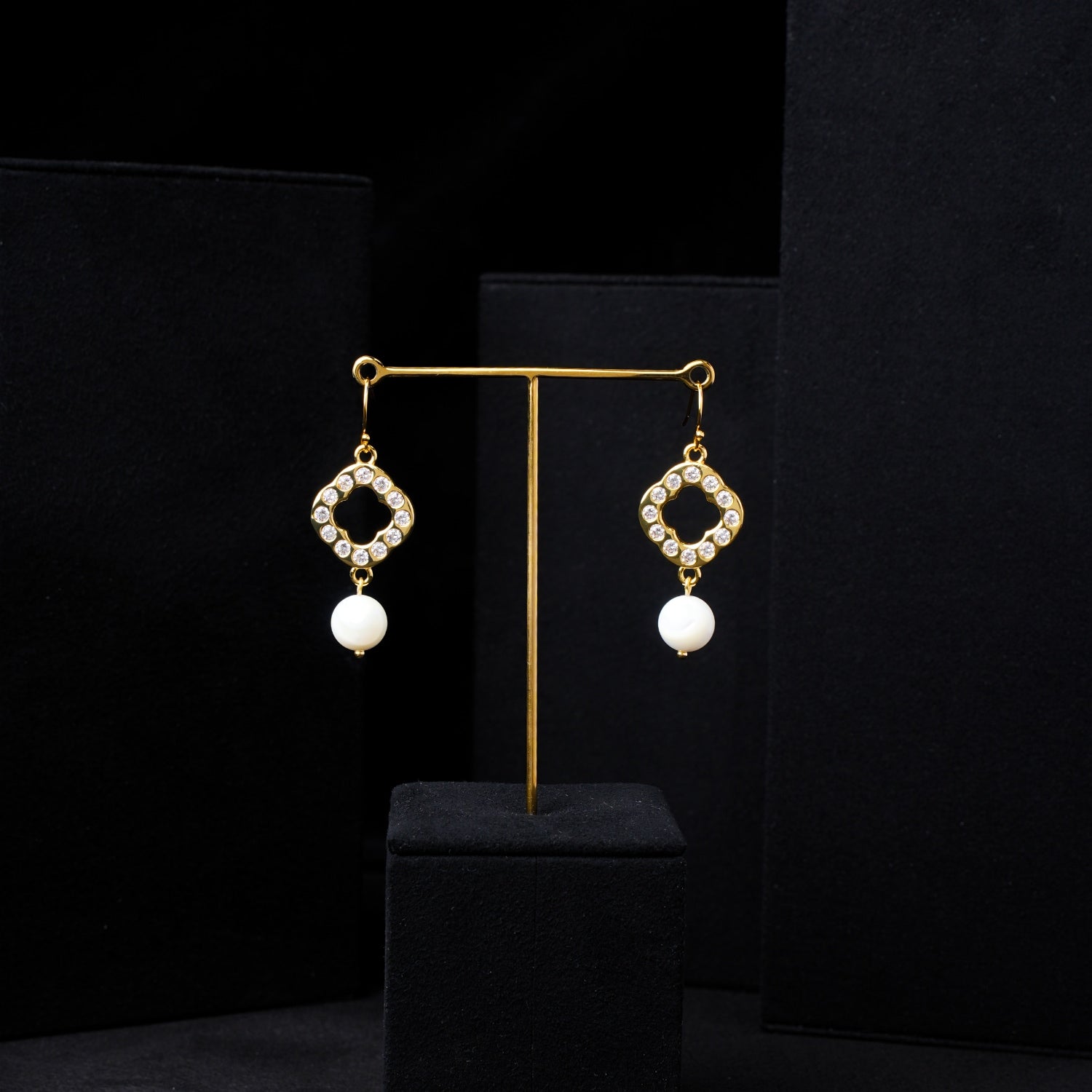 Pair of classic pearl and zirconia gold-plated quatrefoil dangle earrings, elegantly displayed on a minimalist black stand against a layered dark background. Showcasing luxury Nuwa Stone jewelry.