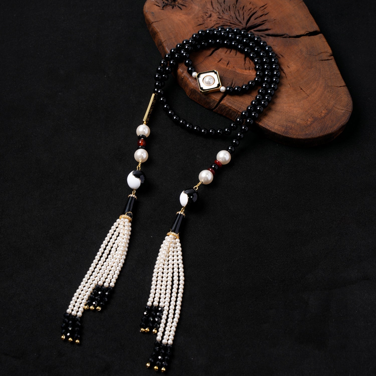 Nuwa Stone's "Grain of the Ocean" multi-strand black onyx and pearl necklace, featuring a central Yin-Yang motif bead, elegant white seed bead tassels, and a gold-plated pearl charm, artfully displayed on a dark wooden board.