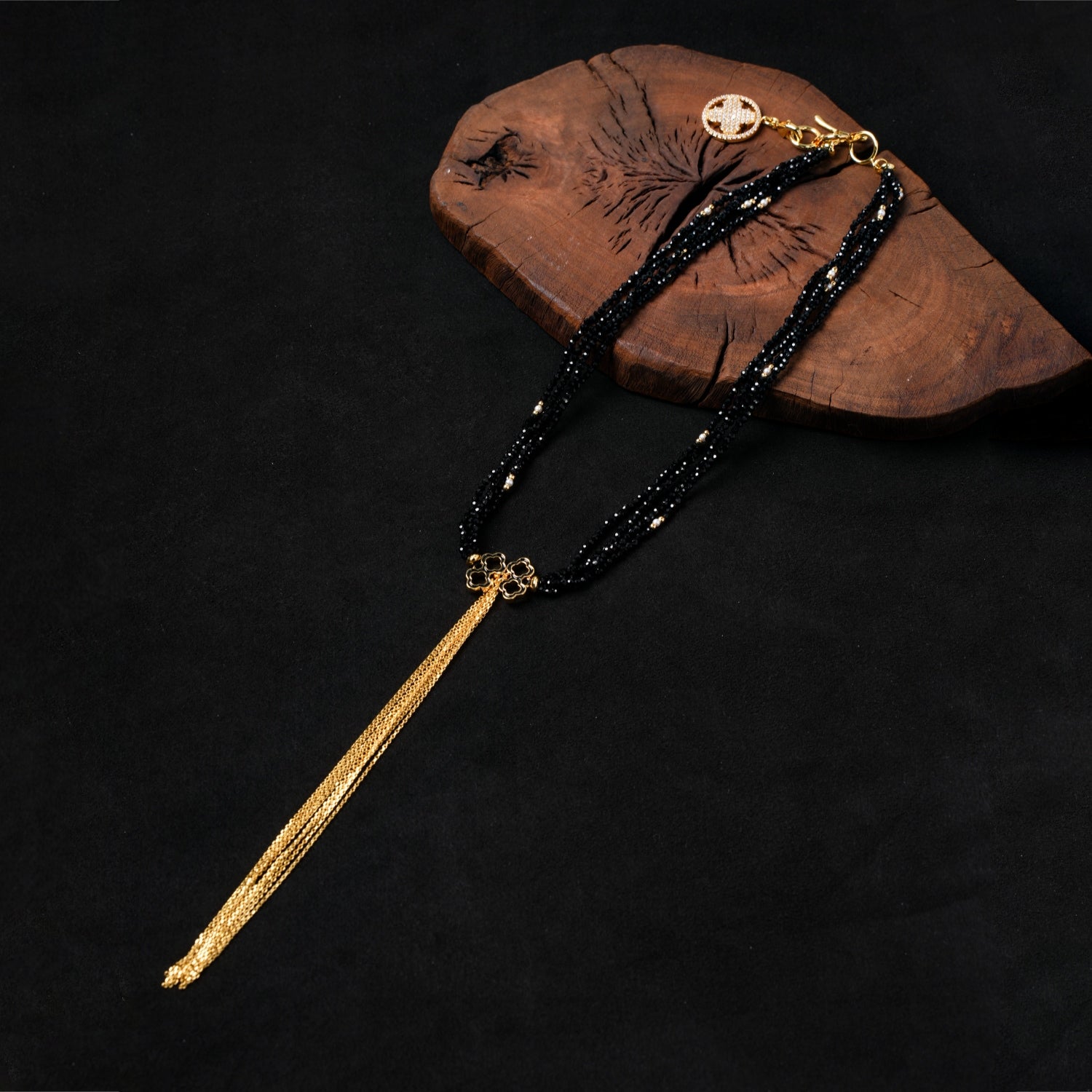 Nuwa Stone's "Ink Begonia Cascading Snow" black spinel and pearl multi-strand necklace with a prominent gold-plated quatrefoil charm and luxurious gold tassel, elegantly displayed on a dark wooden board against a black background.