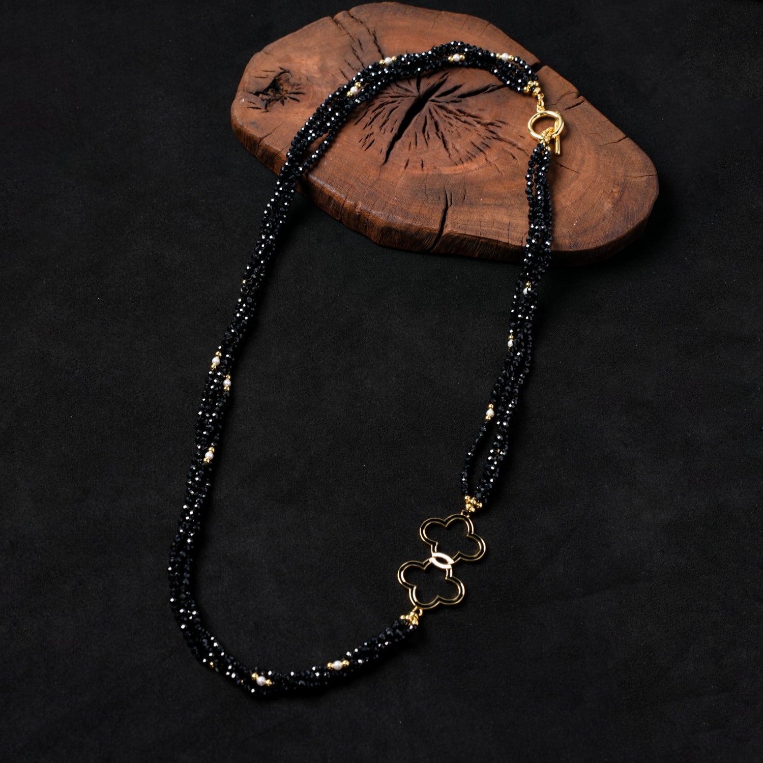 Nuwa Stone's "Nightfall Galaxy Cascade" multi-strand necklace, featuring sparkling black spinel beads, delicate pearls, and a central gold-plated double quatrefoil charm, elegantly draped on a dark wooden board.