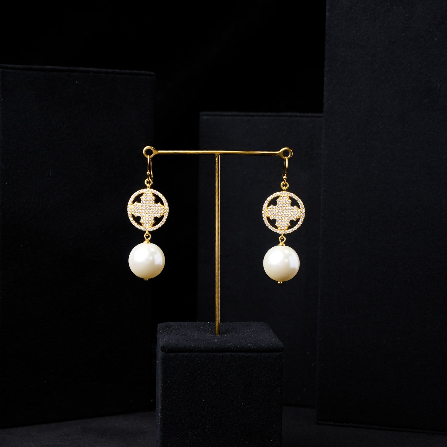 Close-up of elegant pearl and zirconia gold-plated celestial cross dangle earrings on a black display stand. Highlights the lustrous white pearls, sparkling zirconia medallion, and gold setting for Nuwa Stone jewelry.