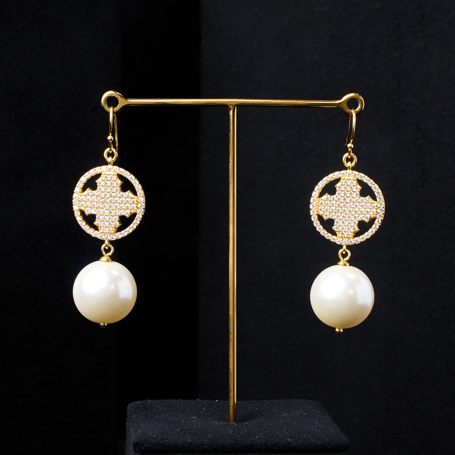 Pair of lustrous pearl and gold-plated sterling silver dangle earrings with a sparkling zirconia celestial cross design, presented on a minimalist black stand against a layered dark background. Showcasing luxury Nuwa Stone jewelry.