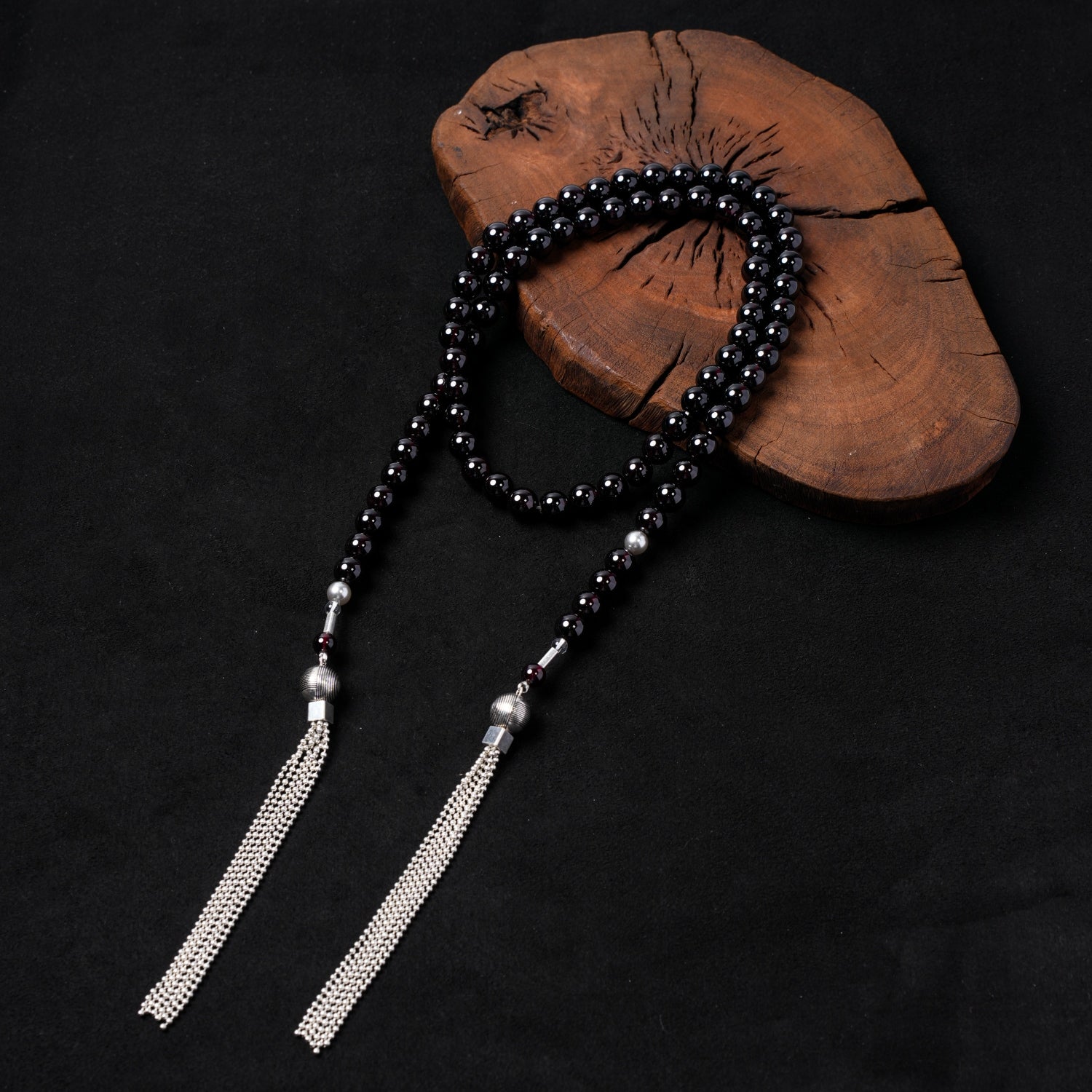 Nuwa Stone's "Twilight Pearl Radiance" long multi-strand necklace, featuring deep red garnet beads, sterling silver accents, black onyx, and two elegant sterling silver tassels, artfully displayed on a dark wooden board.