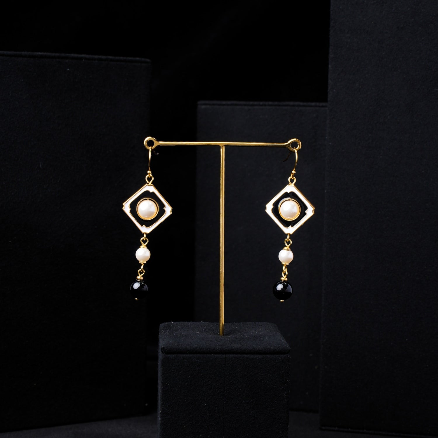 Rear view of Nuwa Stone's Yin Yang Harmony gold-plated dangle earrings on a black display stand. Showcasing the reverse side of the square frames, pearls, and black onyx beads against a dark background.