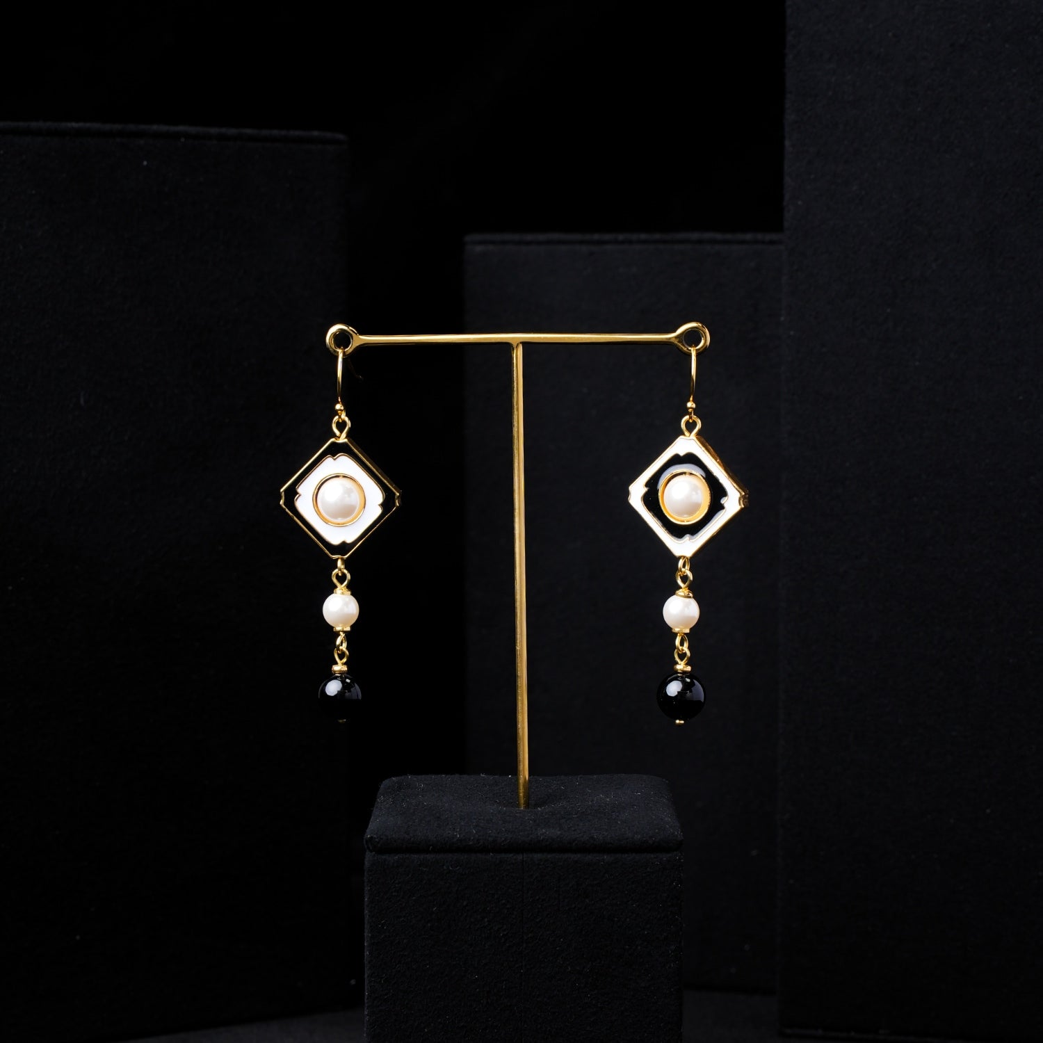 Close-up of Nuwa Stone's Yin Yang Harmony gold-plated dangle earrings, showcasing a square white enamel frame with a central pearl, a smaller pearl, and a black onyx bead. Detailed view highlights the craftsmanship and balance.