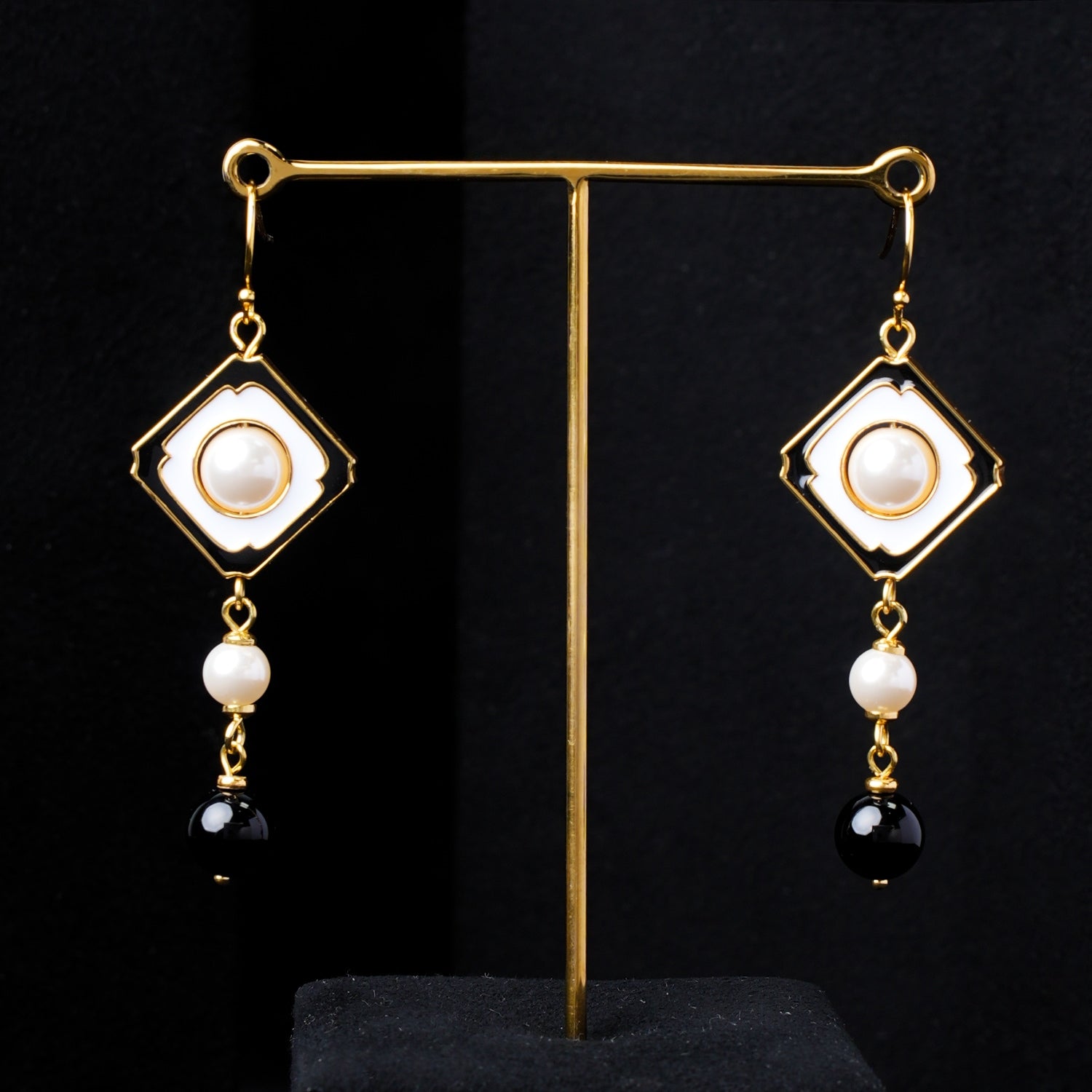 Pair of Nuwa Stone's Yin Yang Harmony gold-plated dangle earrings, featuring square white enamel frames, central pearls, smaller pearls, and black onyx beads, displayed elegantly on a black stand against a dark background.