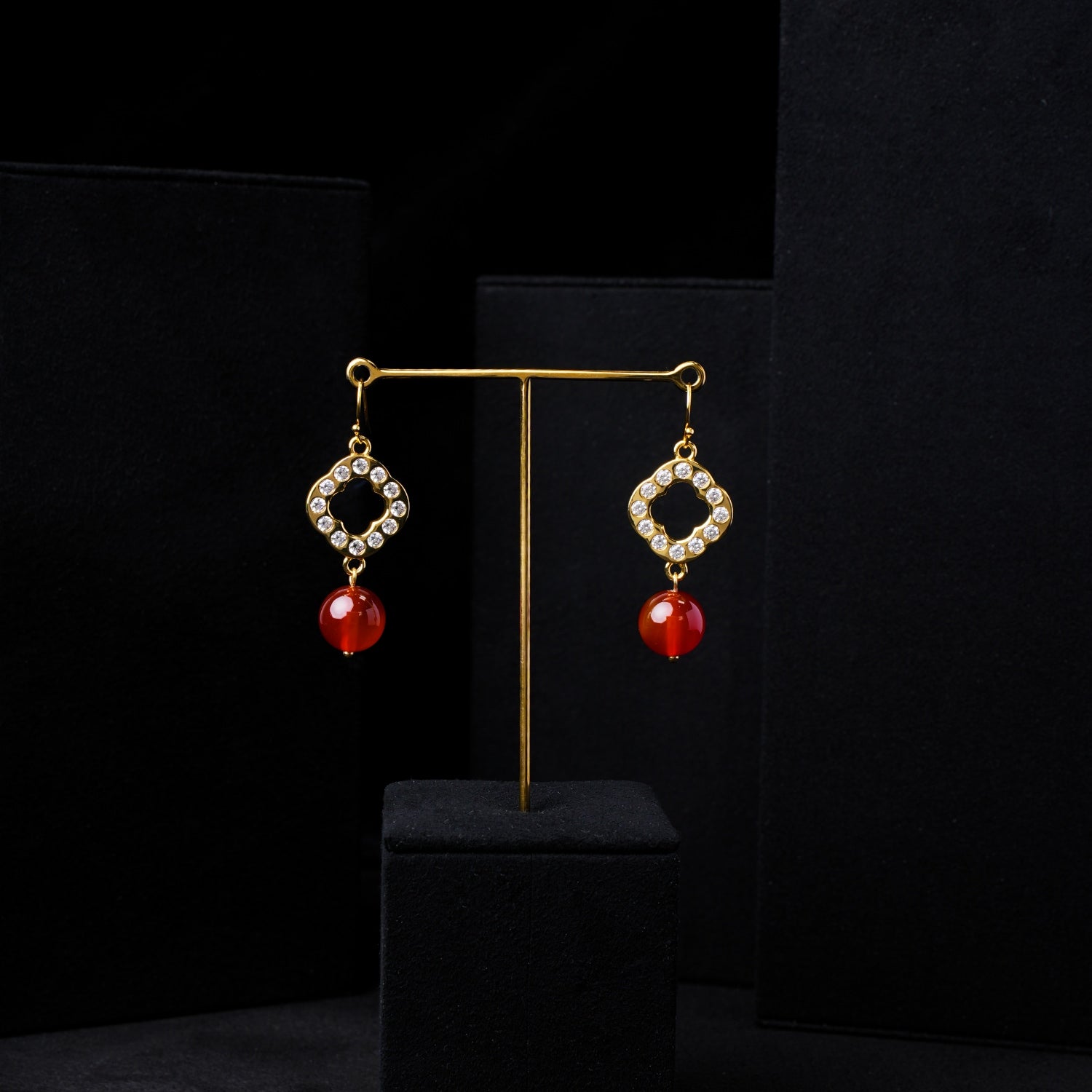 Close-up of elegant red agate and zirconia gold-plated quatrefoil dangle earrings on a black velvet display. Highlights the vibrant red agate beads, sparkling zirconia, and intricate gold setting for Stone World jewelry in Singapore.