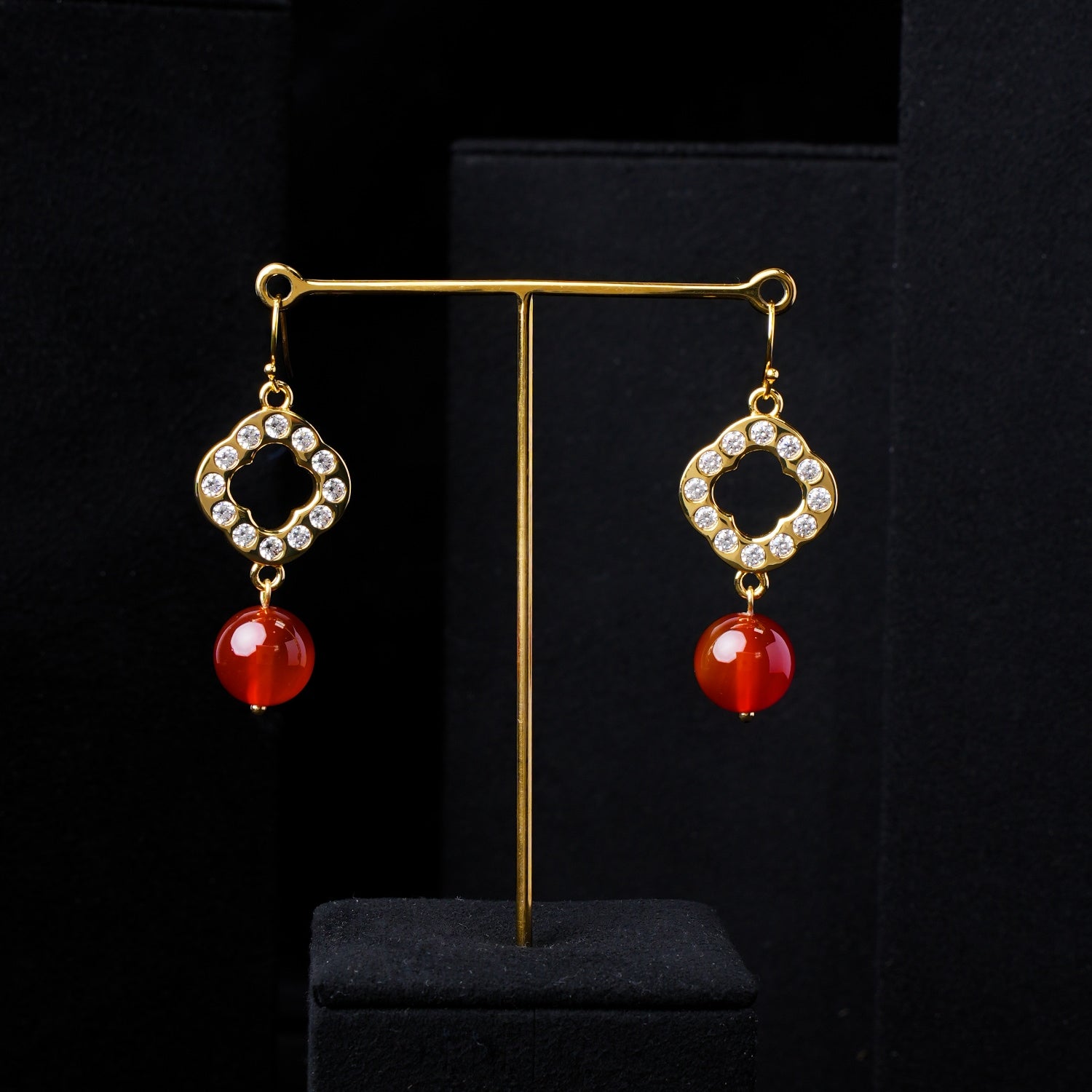 Pair of Red Agate and gold-plated sterling silver dangle earrings with sparkling zirconia, displayed prominently on a minimalist black stand against a sophisticated dark background. Showcasing luxury jewelry from Stone World in Singapore.